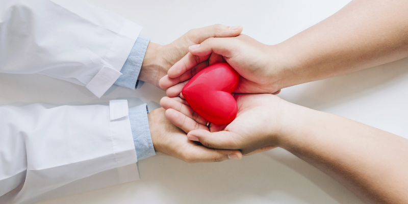 A doctor's hands holding another set of hands which are holding a toy heart. 