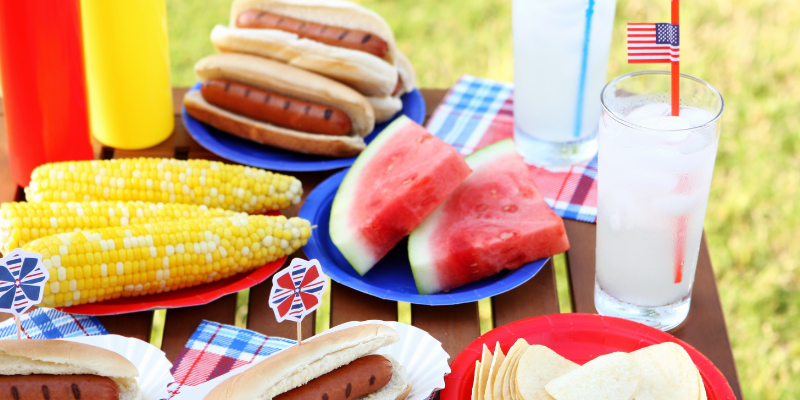 A display of common picnic foods like hot dogs, corn on the cob, watermelon, and chips on a picnic table cloth with Fourth of July accessories. 