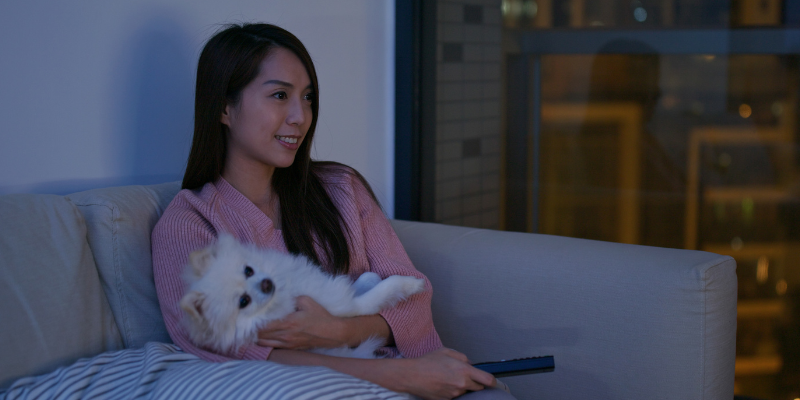 A woman sitting on her couch in a dark room watching TV with her dog in her lap.