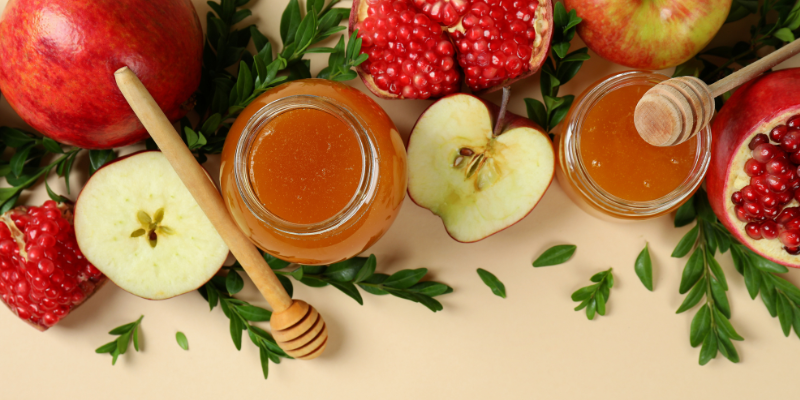 A row of honey jars, apples, and pomegranates on a table.