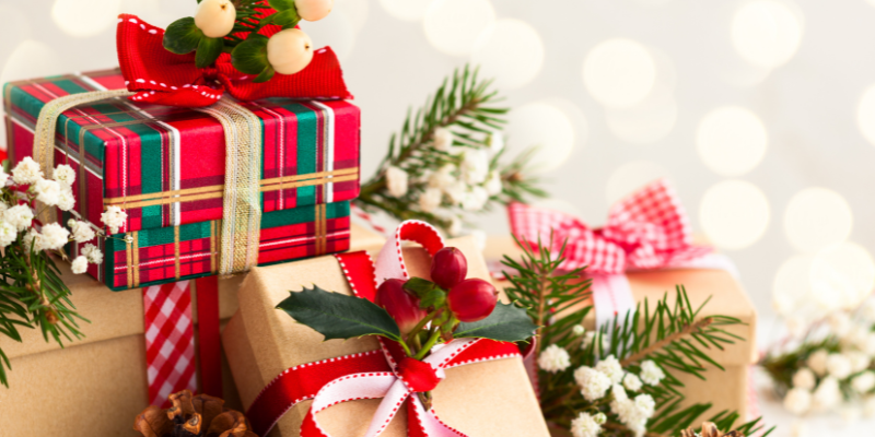 A variety of gifts wrapped with ribbon, string, and decorated with holiday plants in front of a backdrop of Christmas lights.
