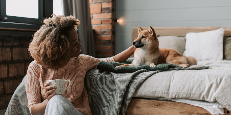 A dog sitting in a bed. A woman sitting on the floor, holding a cup of coffee and petting the dog.
