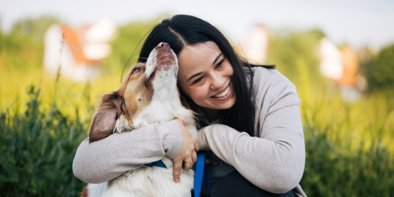 A woman sitting outside while hugging her dog while the dog is looking up to lick her.
